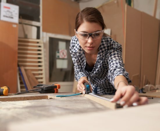 Young,Woman,Woodworker,With,Wooden,Board,Behind,Workbench
